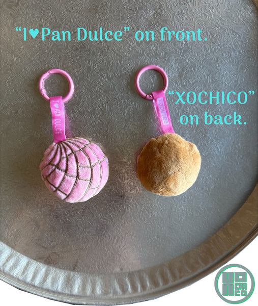 Front and back view of plush concha keychains with 'I❤️ Pan Dulce' and 'Xochico' text on ribbon. Shown on a charola tray.